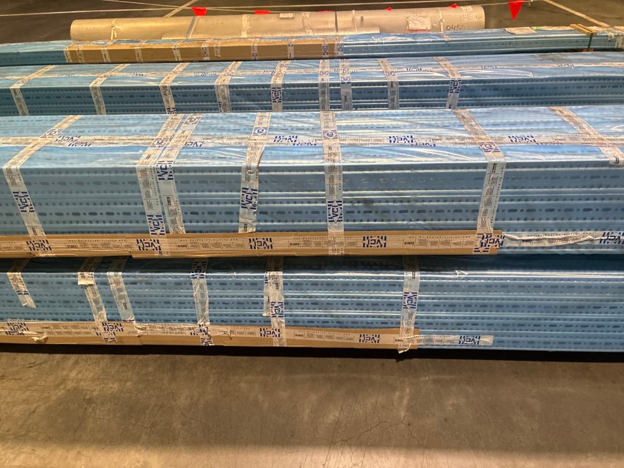 Stacks of blue metal beams wrapped in protective packaging, placed on a warehouse floor.