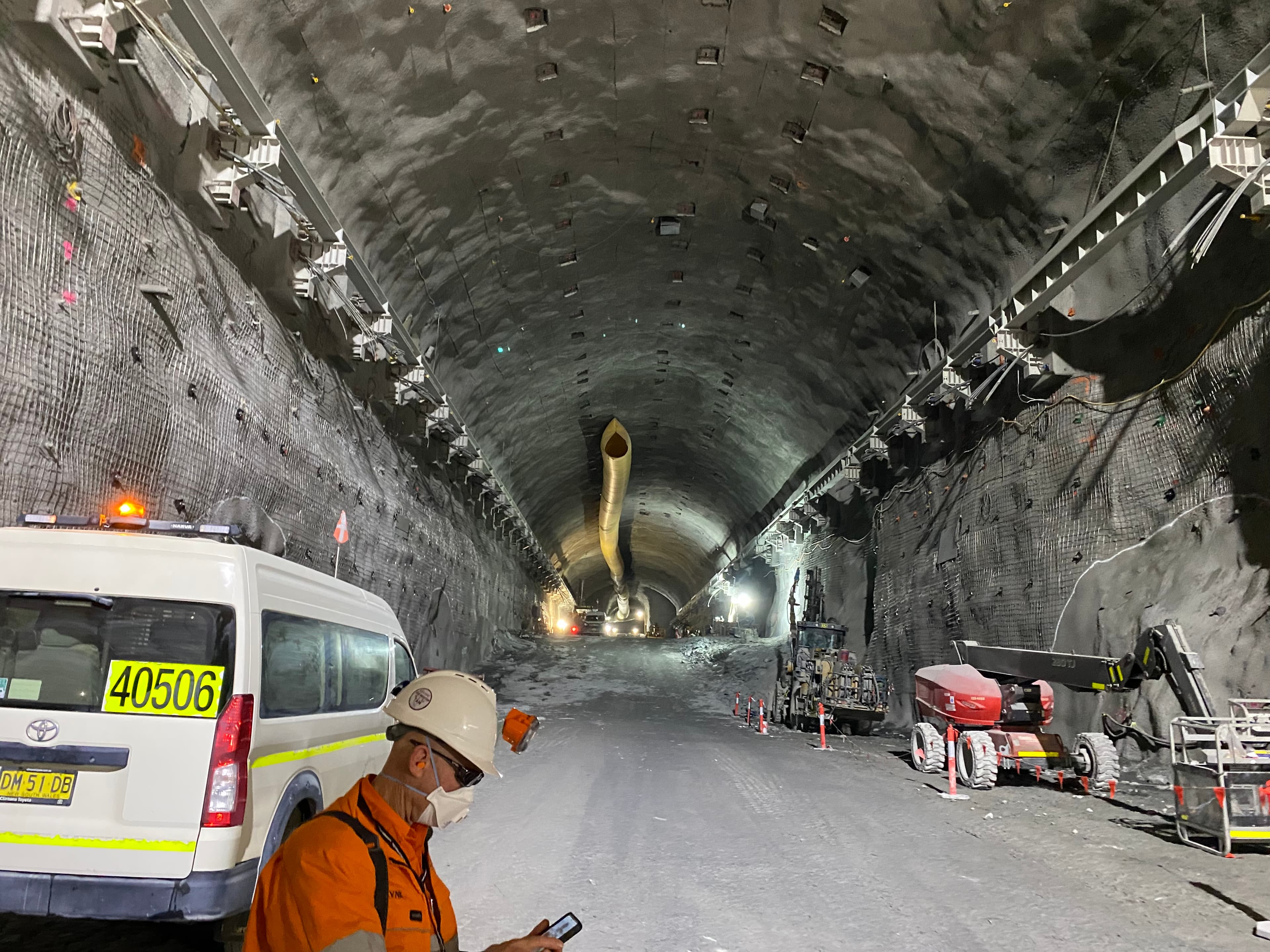 Construction worker in an orange vest and helmet checks phone inside a large, well-lit tunnel with machinery and a ventilation tube overhead.